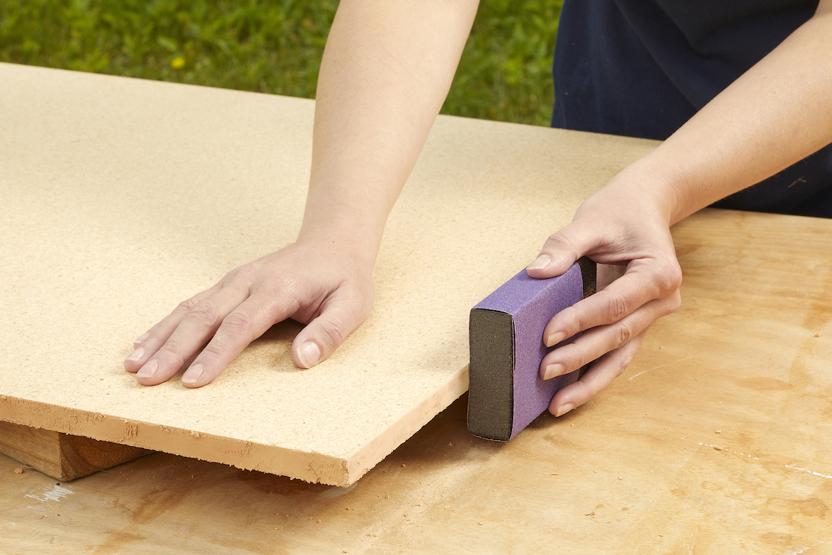 Woman uses a sanding block to sand the edges of particle board.