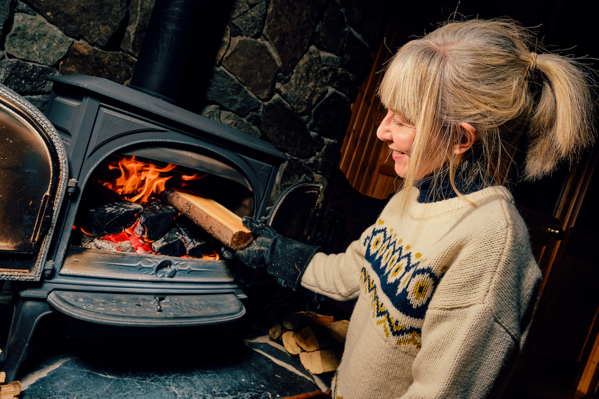 Woman putting a well dried log into a glowing wood burning stove.