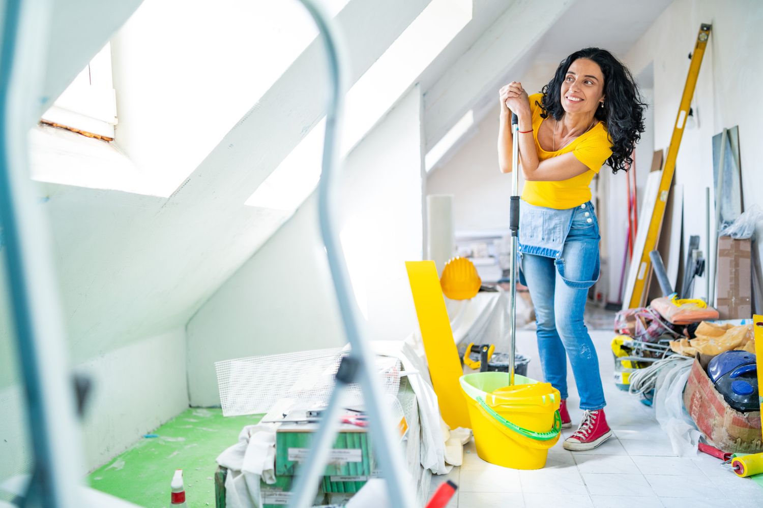 A smiling woman holds a mop while standing in an attic. 