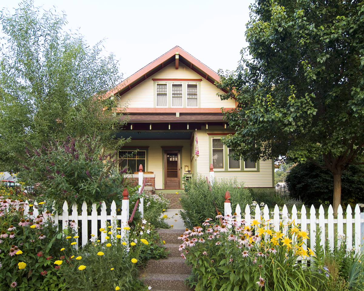 A white picket fence and flowers line the front yard of a bungalow house.