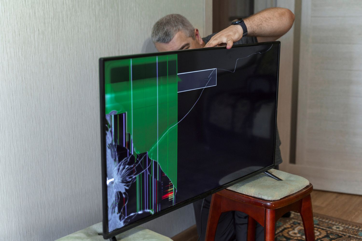 A repair person balances a TV with a broken screen on two stools while they inspect the back of the screen.