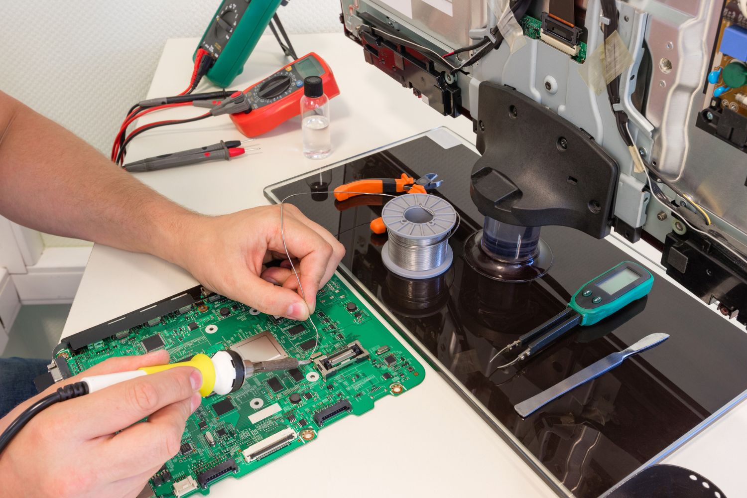 A repair person with only their hands visible uses a soldering tool to repair a motherboard on a broken TV screen.