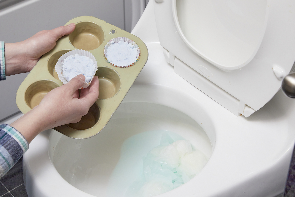 Woman holds muffin tin with DIY drain cleaning tablets near toilet, poised to drop one in.