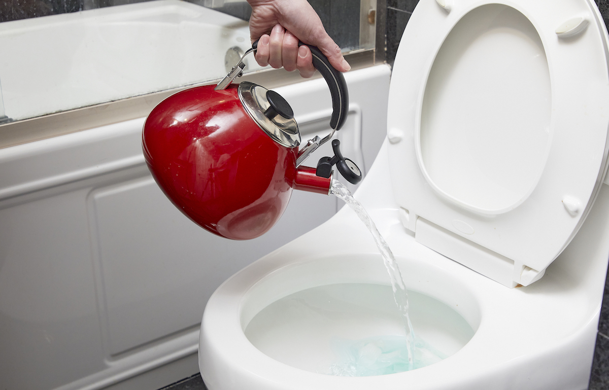 Person holding a red kettle pours hot water into a toilet bowl.