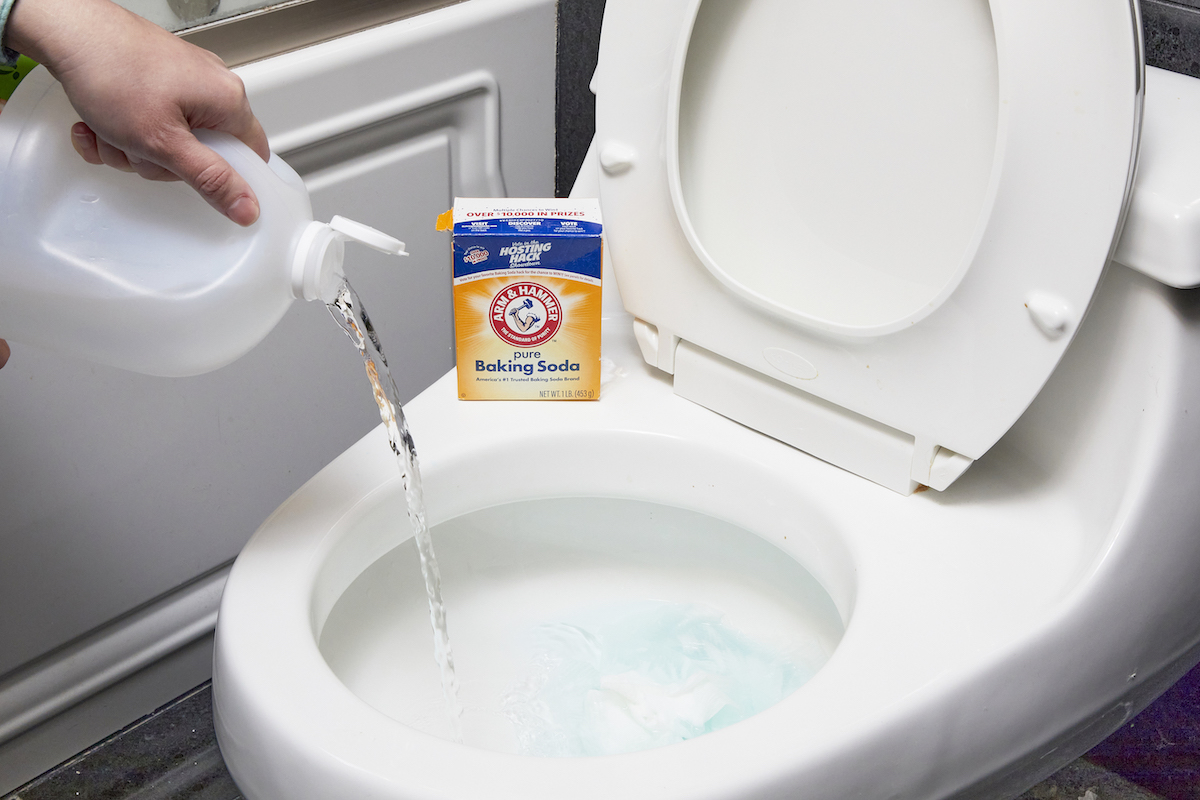 Person pours vinegar from large jug into toilet bowl; box of baking soda sits on toilet bowl. 