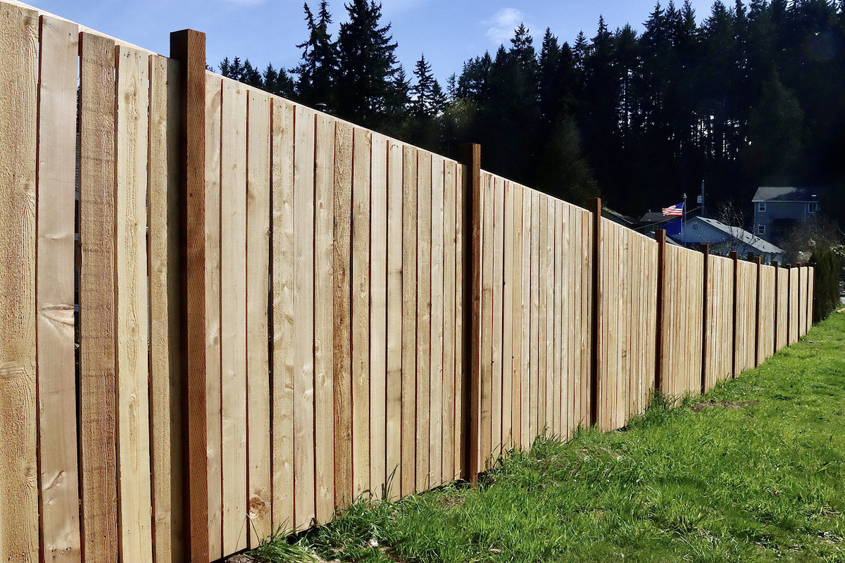 A pine fence lines a backyard in a neighborhood with an American flag and forest in the distance.