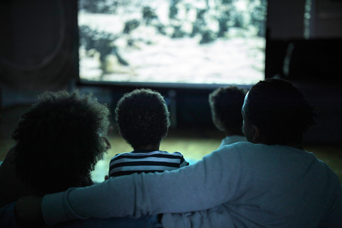 Back view of a family with two children watching an illuminated TV in the dark at home on the couch.