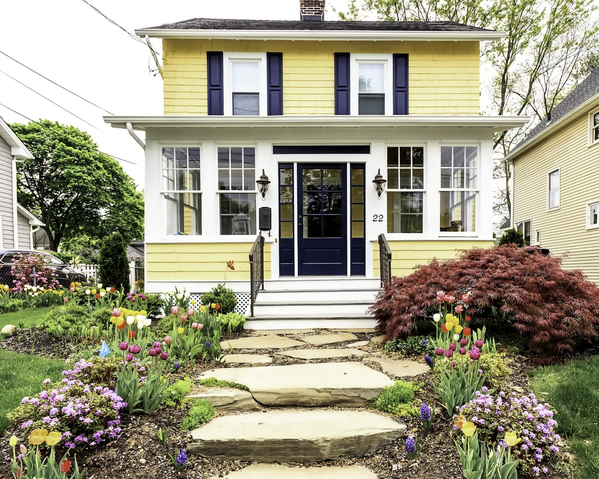 A yellow cottage home with a porch and stone walkway to the navy blue front door.