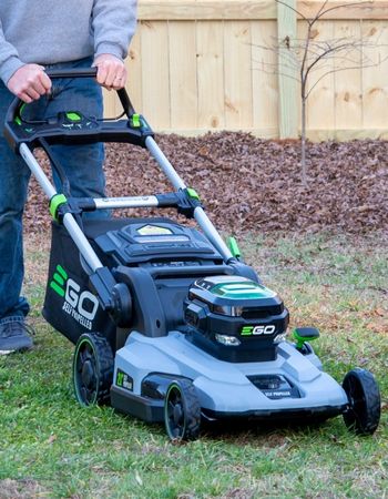 Person pushing Ego lawn mower on grass in front of wood fence