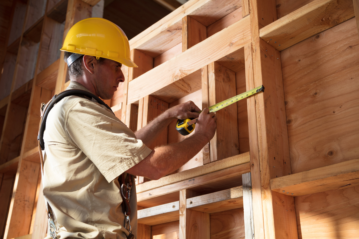 Man wearing construction hat measures space between studs in an open wall. 