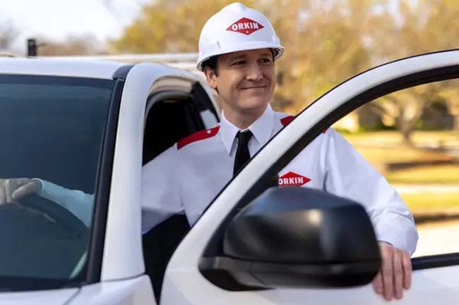 An Orkin employee wearing a company uniform stands by the open door of a white truck and smiles.