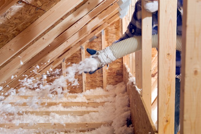 A worker sprays foam insulation onto rafters in an attic.