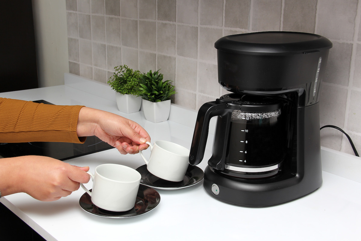 Woman sets two coffee cups next to a coffee maker with a carafe full of coffee.