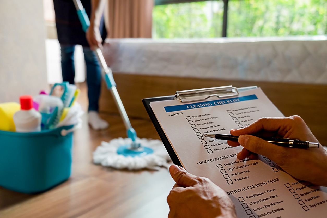 woman's hands checking a checklist on a clipboard while another woman mops a bedroom floor