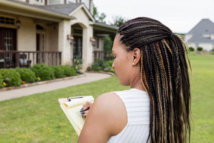 a-woman-with-long-braids-looks-down-at-her-clipboard-as-she-stands-in-the-yard-of-a-tan-house