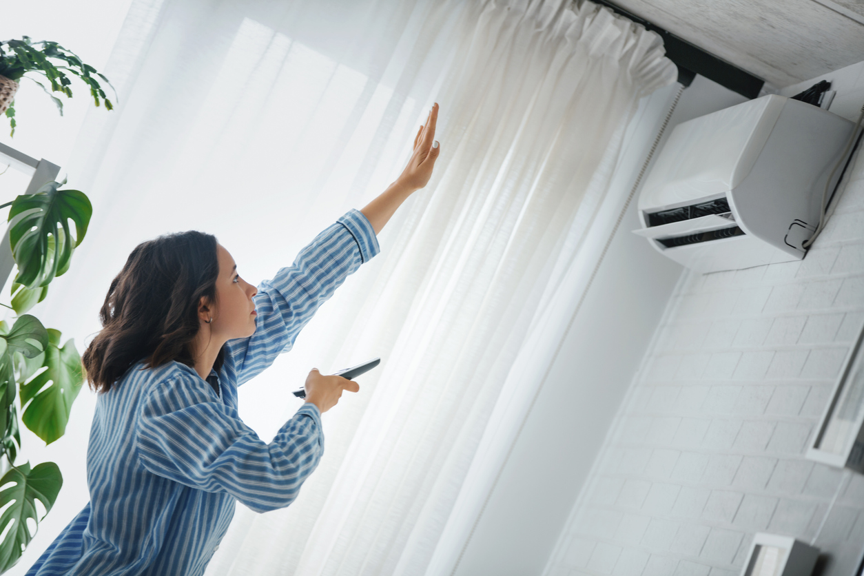 woman in modern bright living room near white curtained window turns on air conditioning mounted on wall