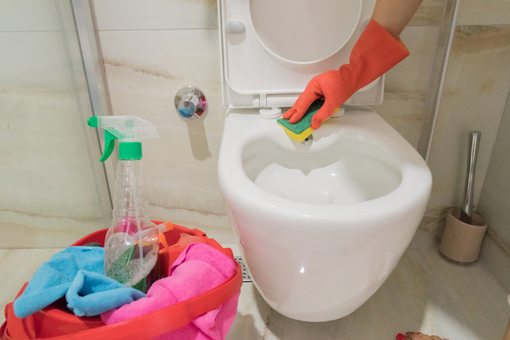 hand-in-organge-glove-using-sponge-to-clean-toilet-with-pink-basket-of-cleaning-supplies-nearby-in-beige-bathroom