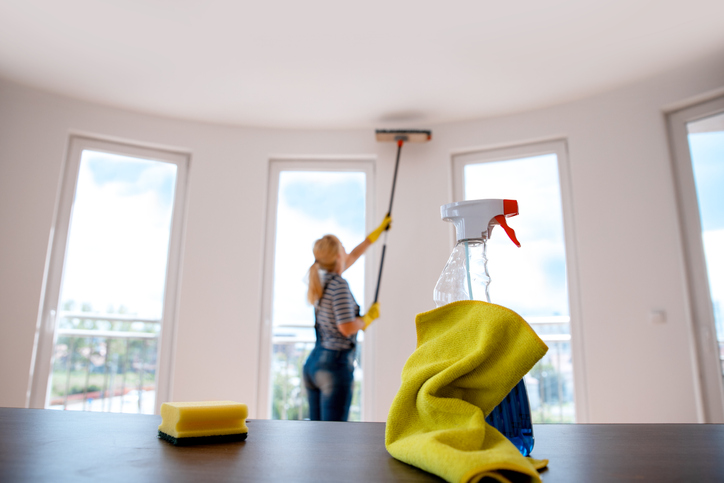 window-cleaner-bottle-rag-and-sponge-on-table-with-blonde-woman-in-background-cleaning-wall-and-windows-with-long-handled-sweeper