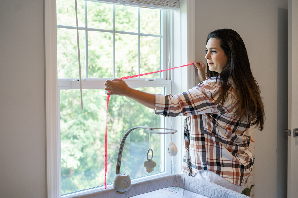 pregnant woman measuring window behind crib in baby room