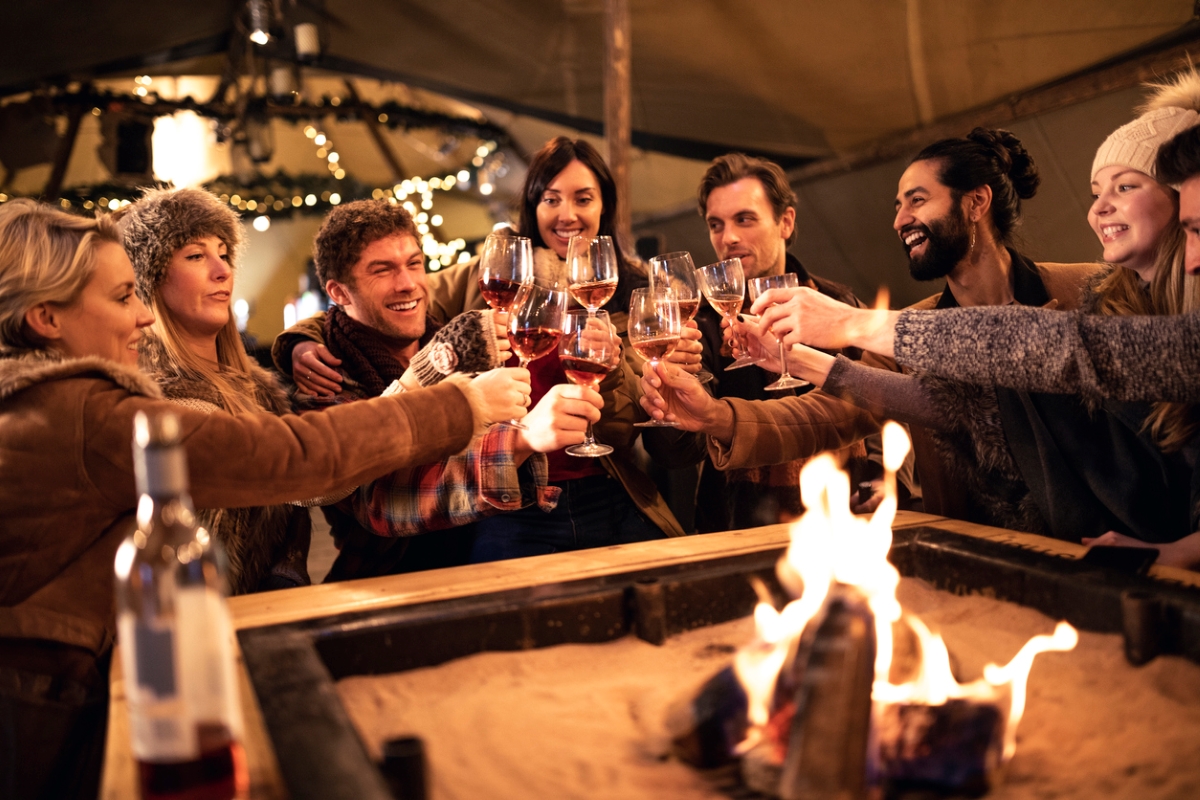 A group of people are holding wine glasses around a fire pit underneath a large canopy.