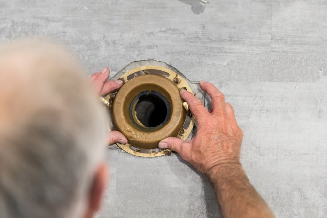 Overhead view of a man setting a wax ring around the toilet pipe opening on a tile floor.