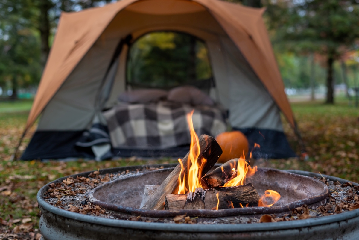 A fire pit is burning in front of camping tent with pillows and blankets inside.