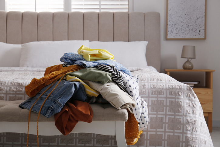 a-messy-pile-of-folded-clothes-sits-on-a-white-bench-at-the-end-of-a-bed-with-a-beige-headboard-and-bedside-table