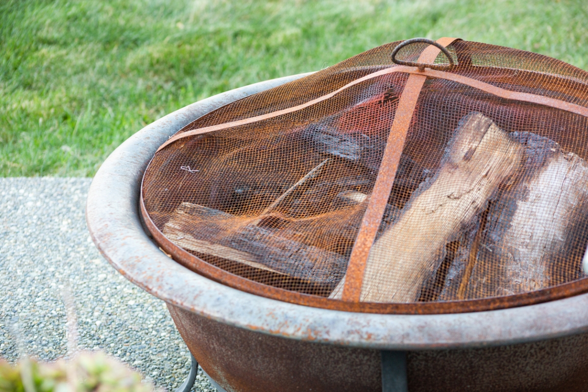 A fire pit with wood inside is covered with a metal mesh screen cover.