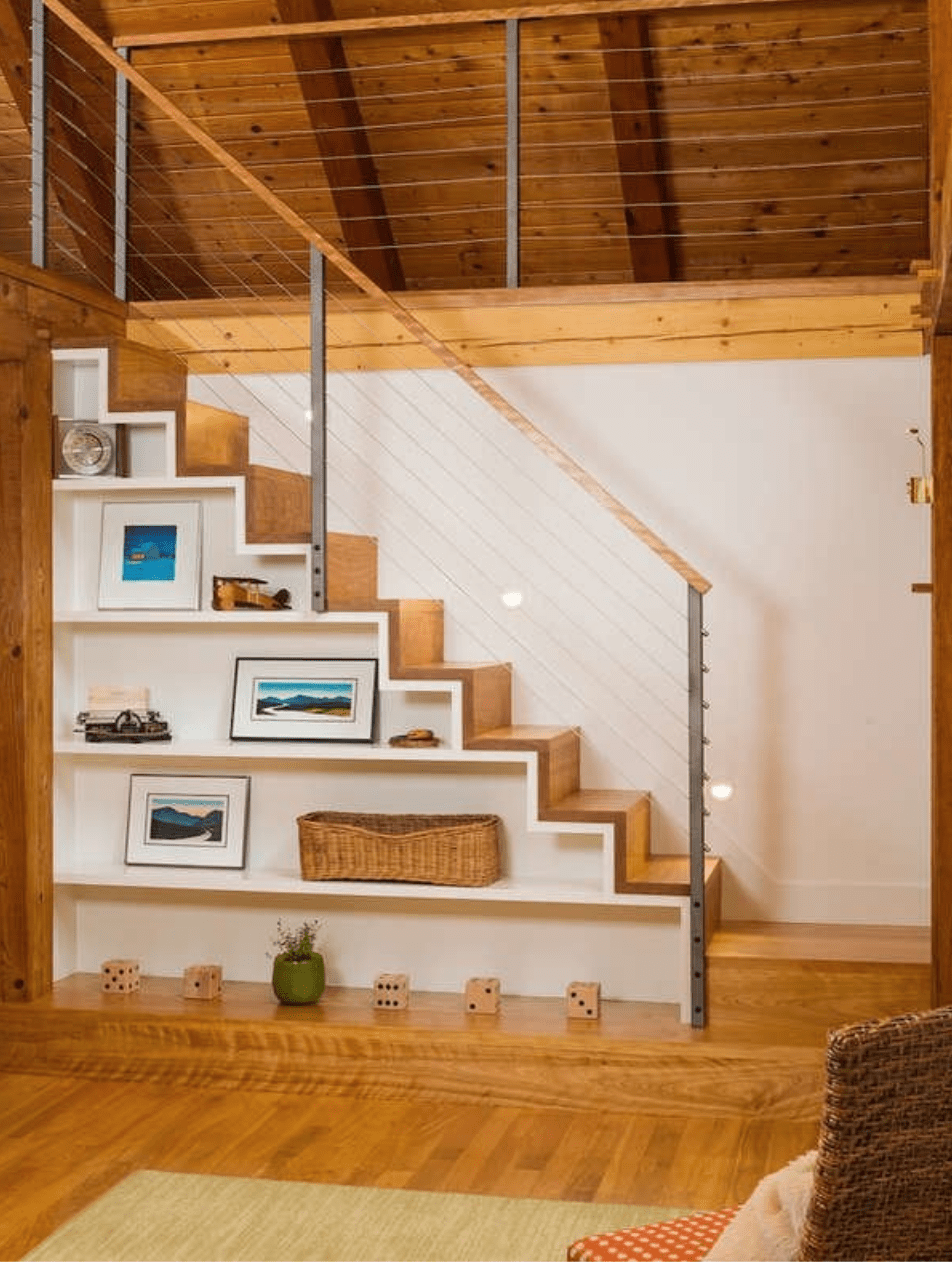 light wood staircase leading into attic space with wood beam ceilings with shelves underneath staircase displaying framed artwork