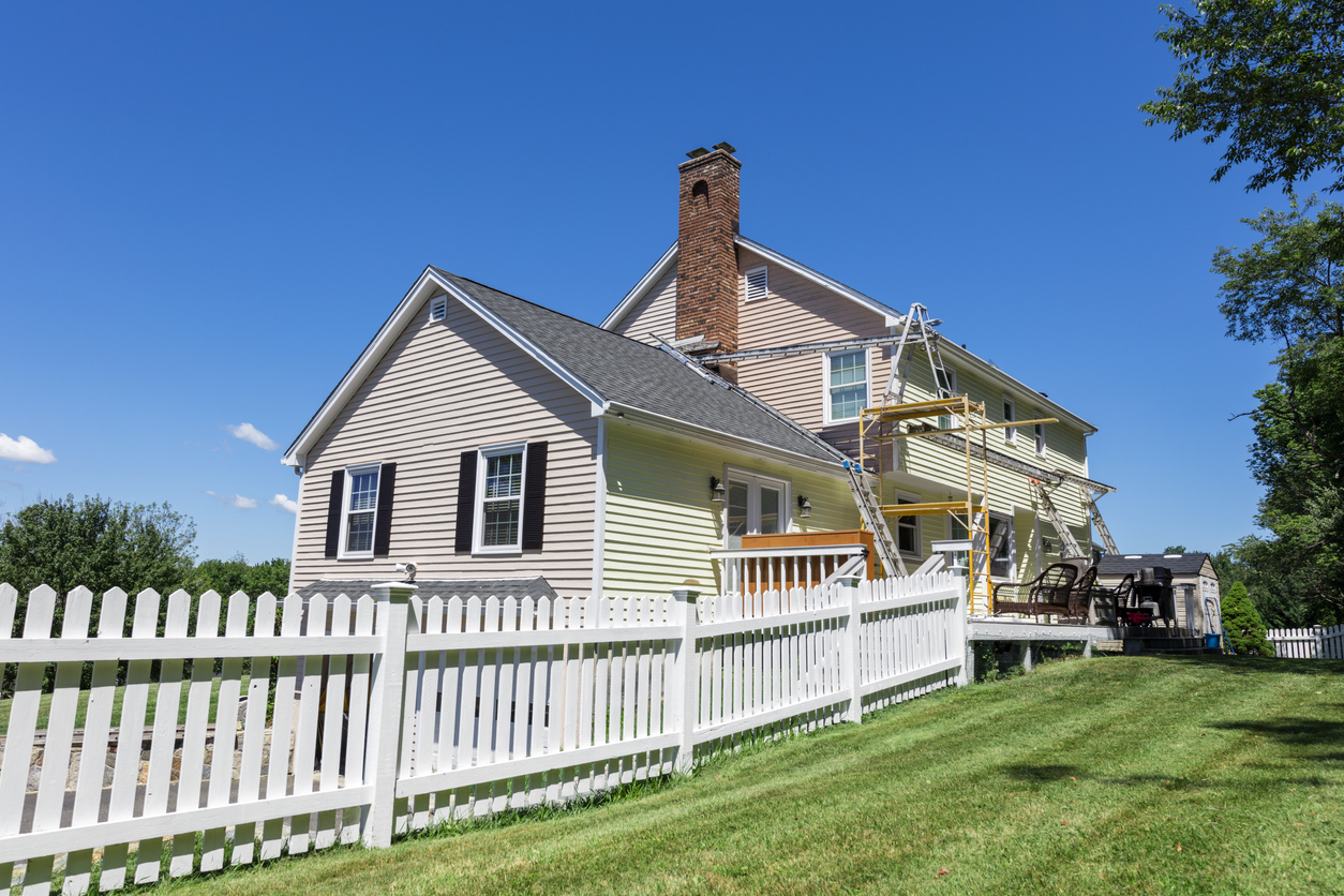 large house on green lawn with one side painted beige and the other yellow with ladders set up indicating a paint job