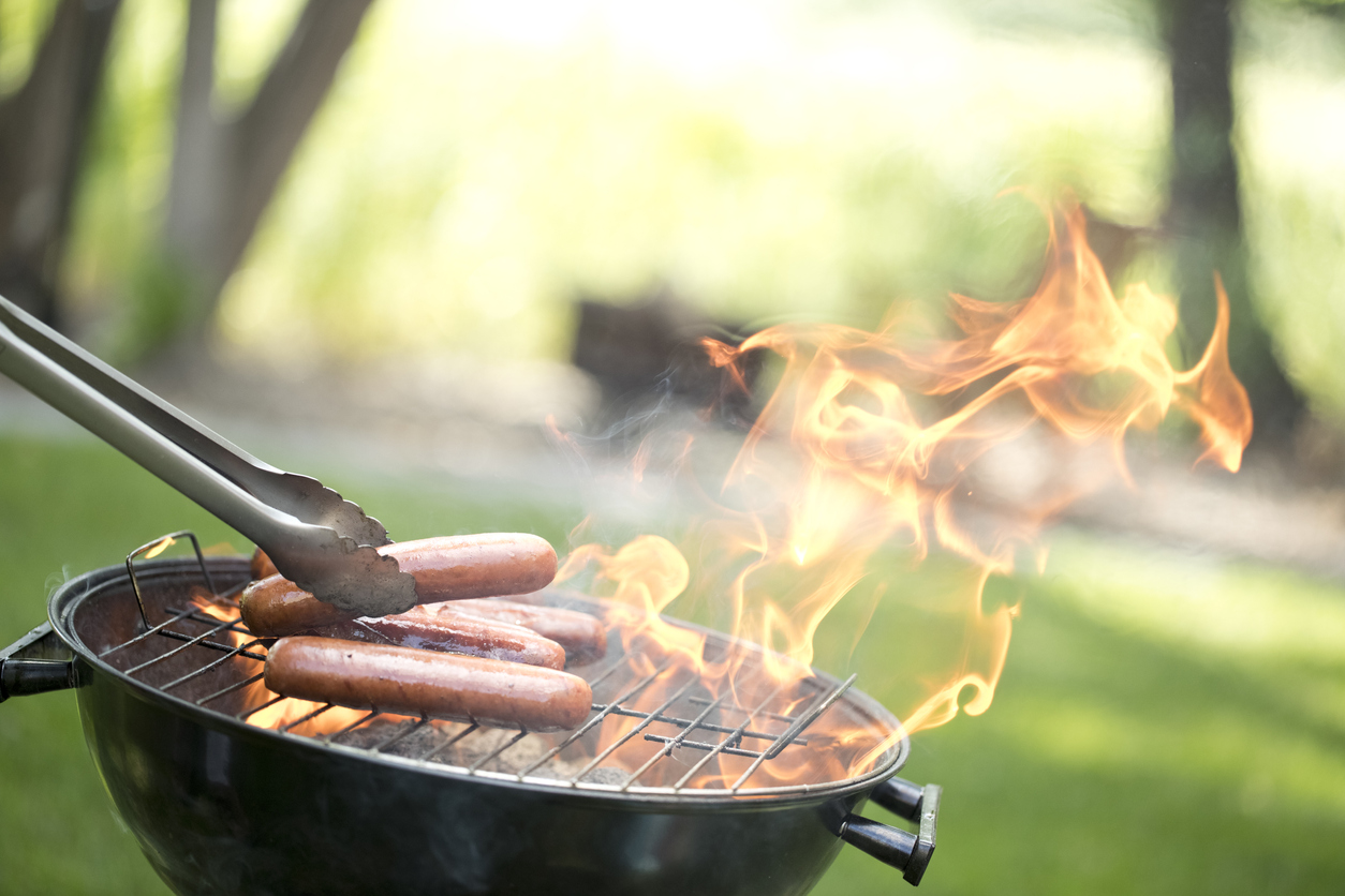 round grill outside on green lawn with tongs placing hot dogs on high open flame