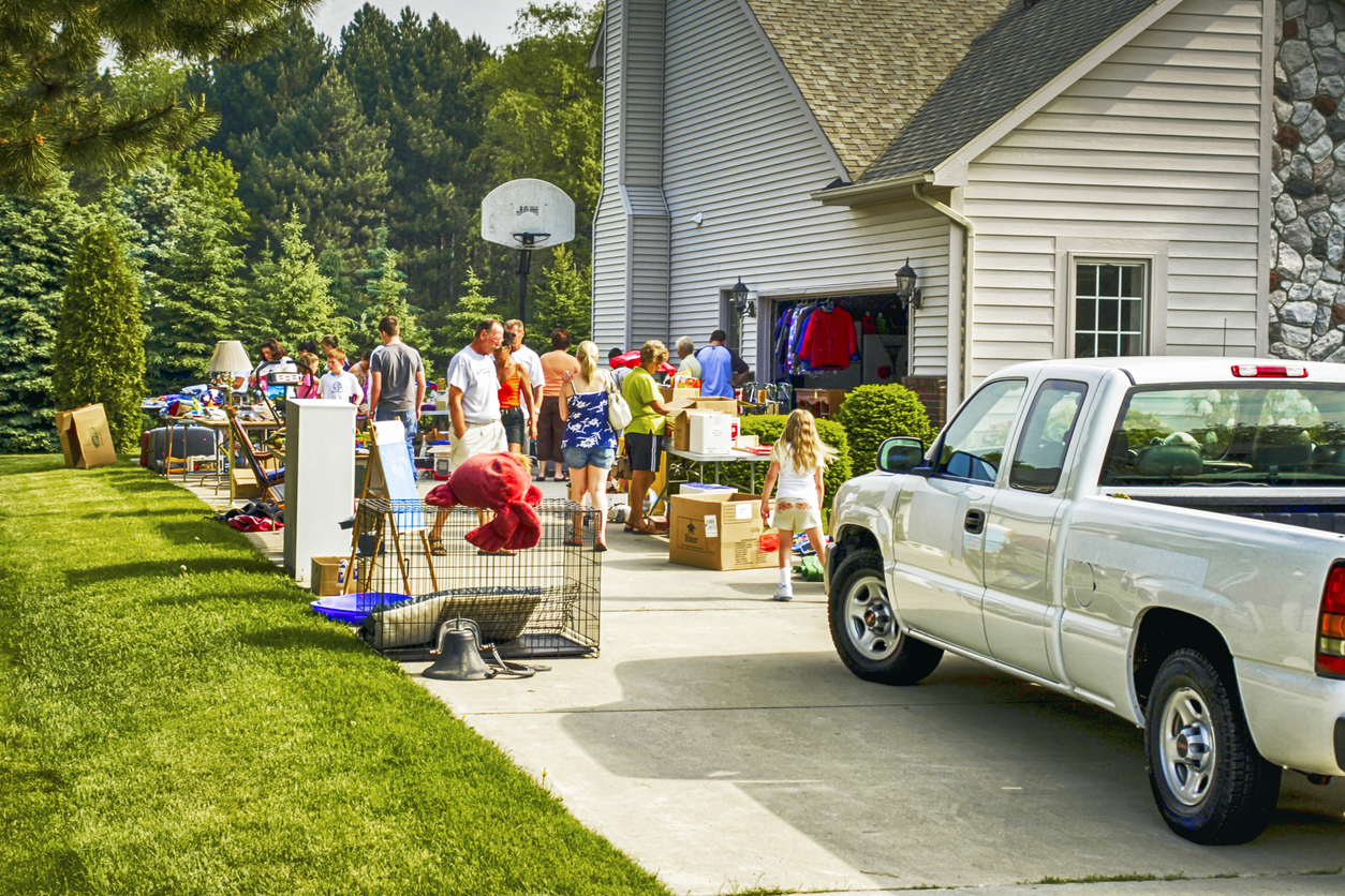 driveway with pickup truck parked and tables set up for yard sale and groups of people shopping outside large house