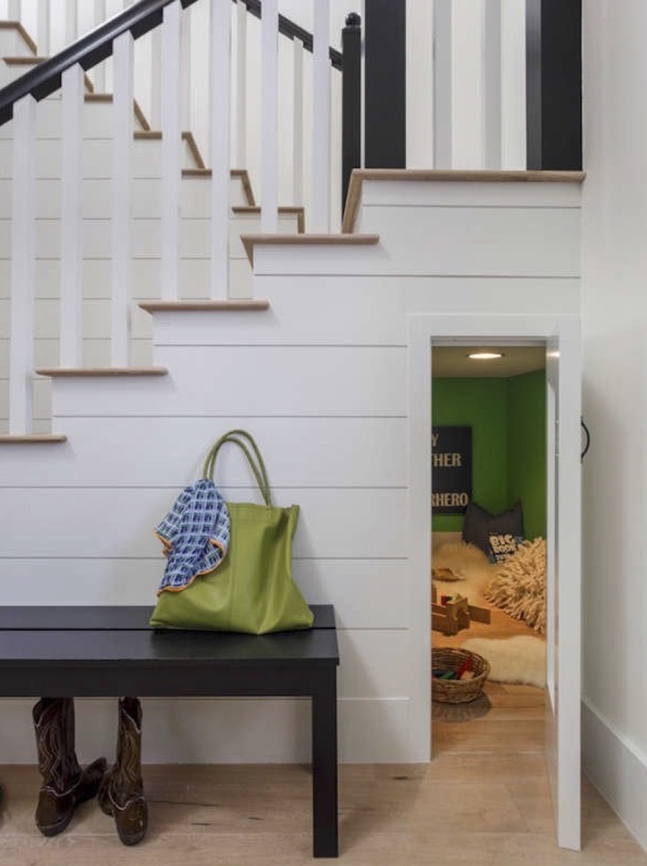 view of white paneling beneath a staircase with cowboy boots beneath a bench and a small open door to a crawl space filled with pillows, carpet, and toys