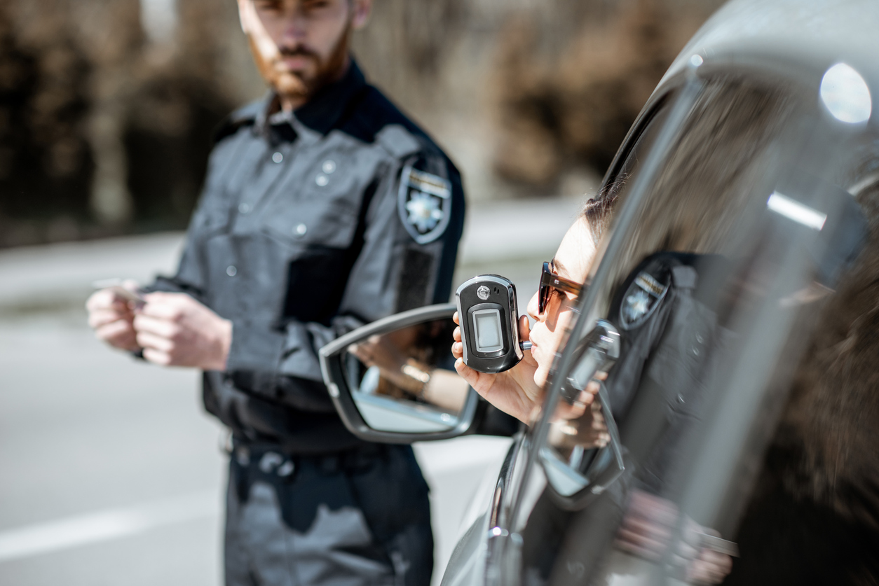 view from behind left side of black car as woman driver takes breathalyzer test leaning out the window and blurry police officer watches at a distance