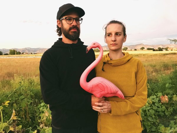 a-young-man-and-woman-in-a-field-hold-a-plastic-pink-flamingo-in-the-same-post-as-the-painting-American-Gothic