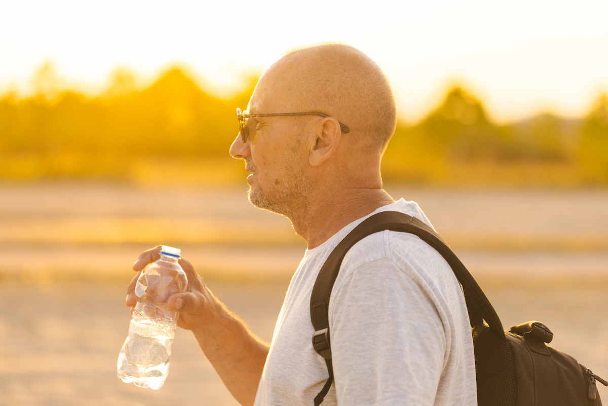 A bald man with sunglasses drinks from a plastic water bottle while sun sets in background.