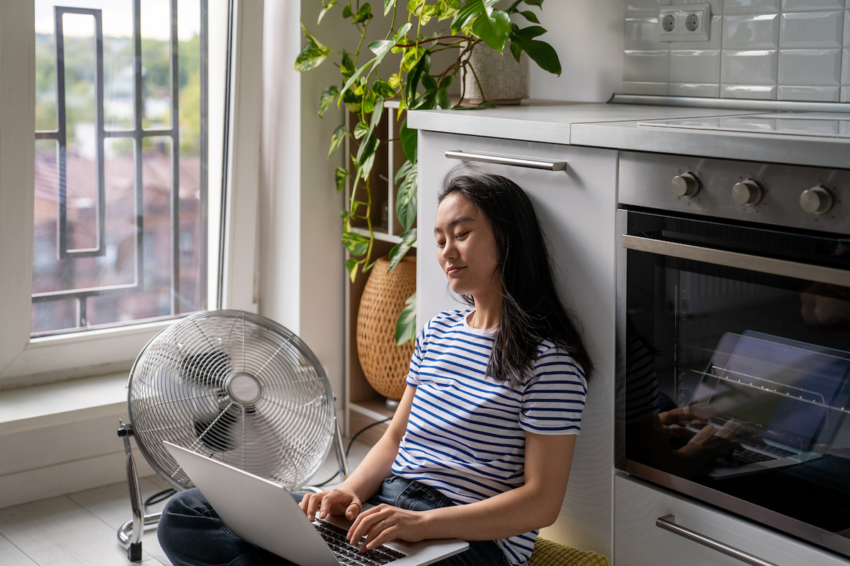 Young Asian woman sitting on the floor in the kitchen, typing on laptop next to an electric fan. 