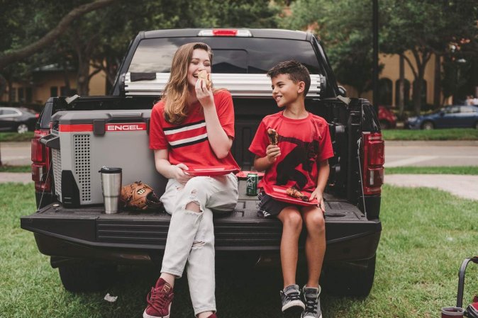 Two people sitting on the tailgate of a truck eating and drinking next to the best electric cooler option