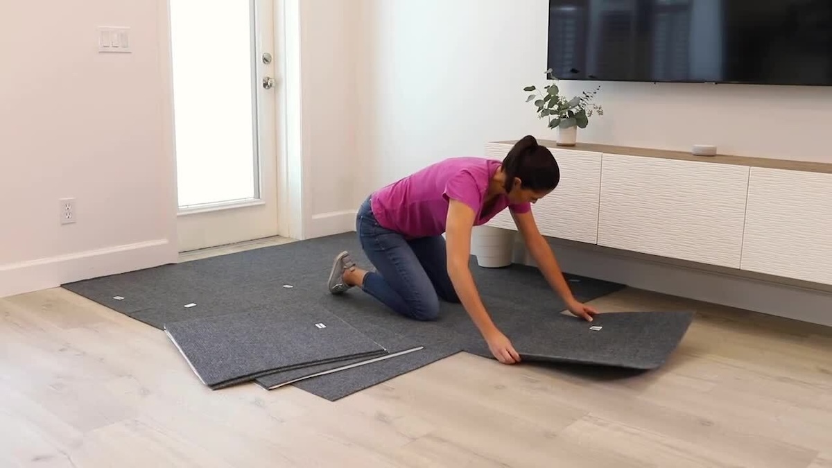 Woman assembles carpet tiles on a floor, with a flat-screen tv on the wall in the background.