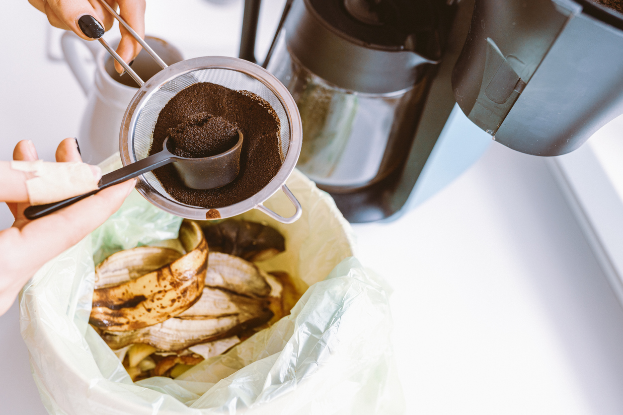 Old coffee grounds being spooned into a compost container next to a coffee maker.