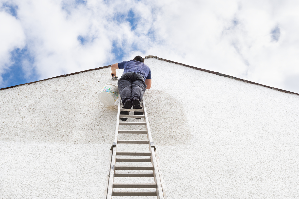 High quality photo of a man up high on a ladder painting a white pebbledash wall on the gable end of a house.