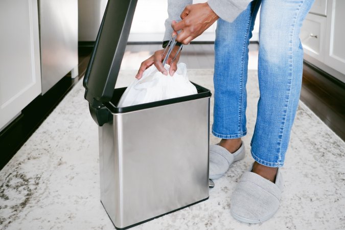 A woman wearing jeans and slippers pulls a full bag of trash from a waste basket.