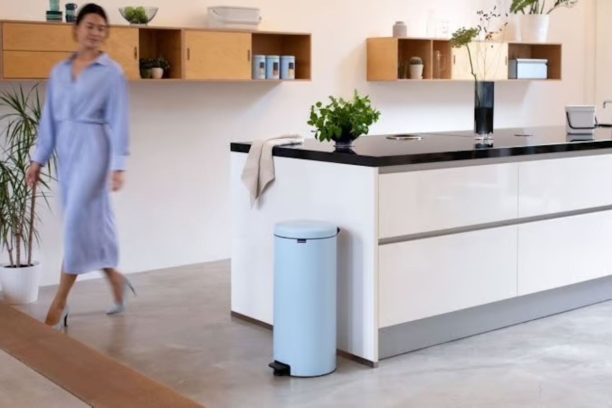 A woman in a kitchen walks past a light blue  5- to 8-Gallon Trash Can. 