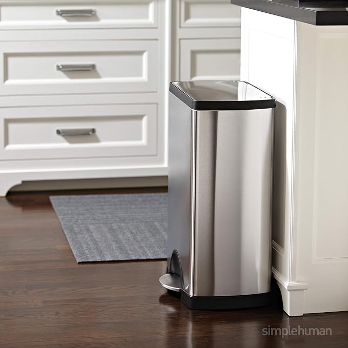 A steel 10- to 20-Gallon trash can stands in a kitchen with white cabinets. 