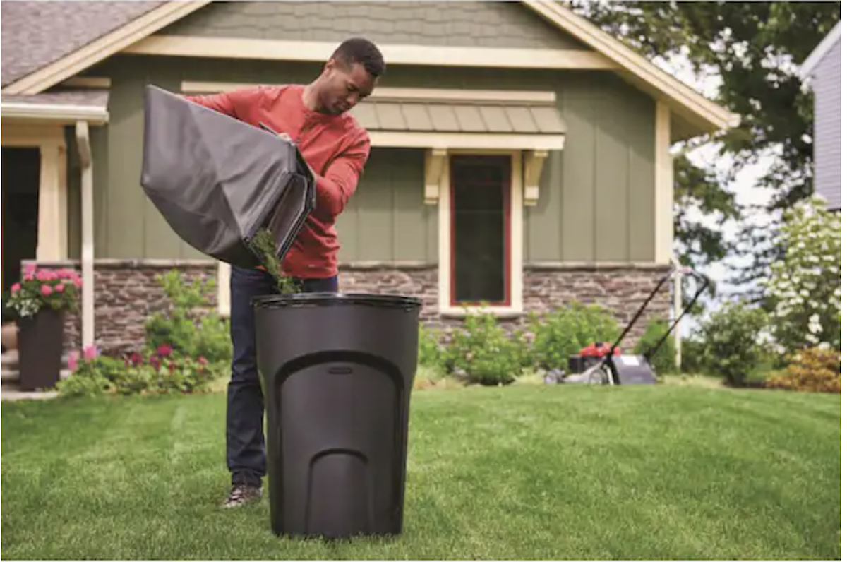 A man in a red shirt pours lawn clippings into a black 30- to 48-Gallon trash can in his front yard. 