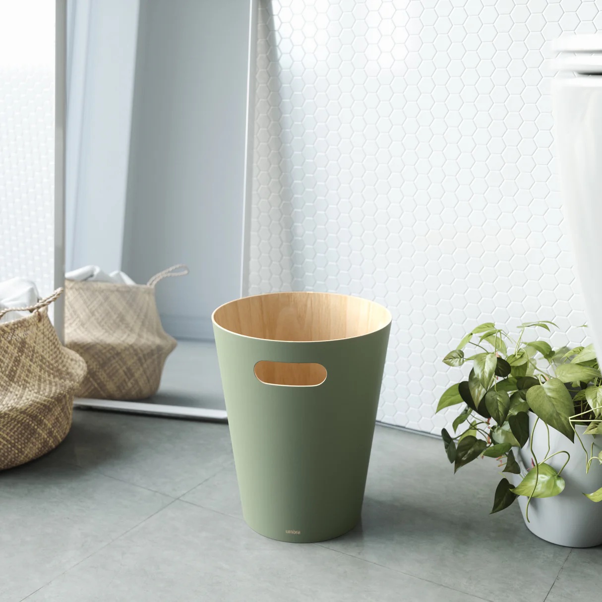 Umbra Woodrow trash can in spruce green near a floor-length mirror and a plant in a bedroom.