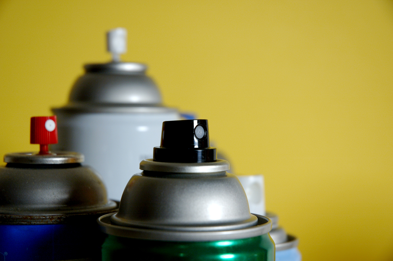 A group of four uncapped spray paint can nozzles in front of a yellow background.