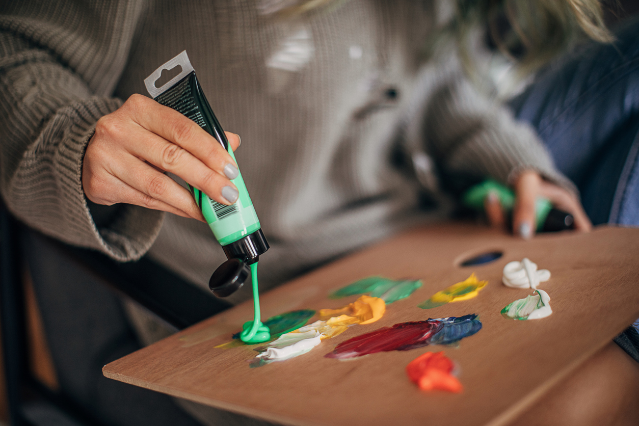 A tube of light-green acrylic paint being squeezed out onto a palette by a woman wearing a sweater.