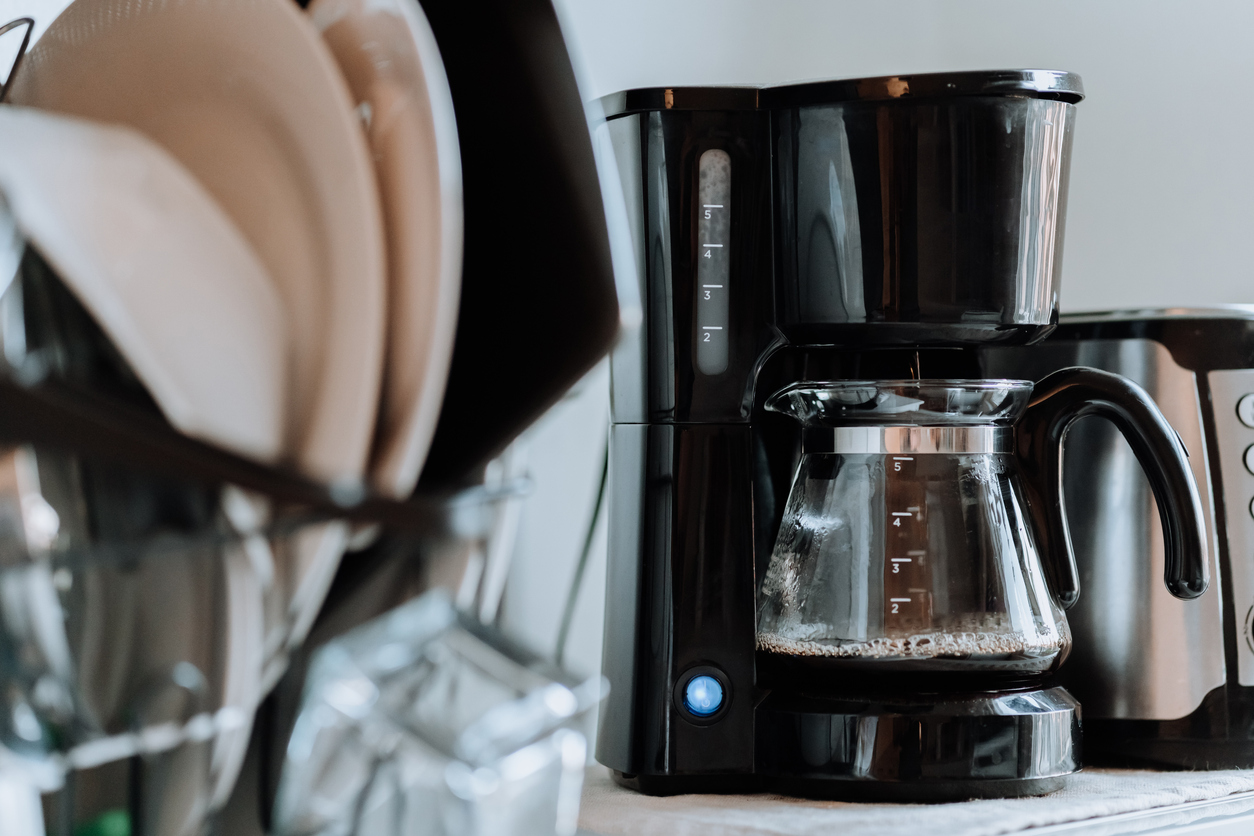 A coffee maker sits on the counter full of coffee behind a rack of clean dishes.