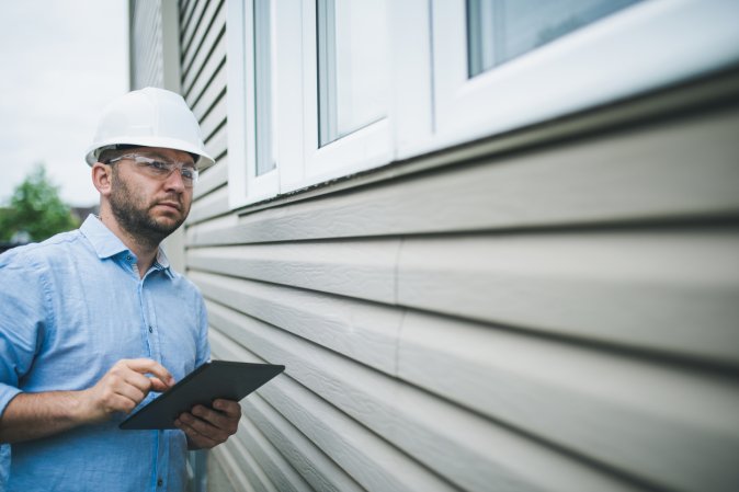 A home inspector with a clipboard in a hard hat looks at the side of a house.